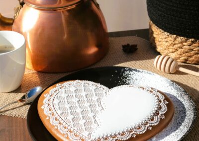 Biscuit décoré en forme de cœur sur une assiette noire, entouré d'une théière en cuivre, d'une tasse de café et d'épices pour une ambiance chaleureuse.