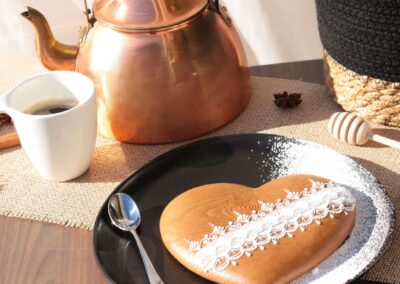 Biscuit en forme de cœur avec texture effet bois et motifs en glaçage blanc, servi sur une assiette noire avec une tasse de café et une théière en cuivre en arrière-plan