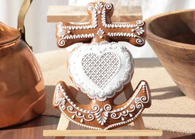 Biscuit décoré en forme de croix camarguaise avec un cœur, magnifiquement orné de motifs en glaçage blanc, présenté sur un chevalet devant une théière en cuivre.