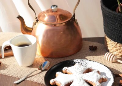 Biscuit en forme de croix d'Occitanie décoré avec du glaçage blanc, posé sur une assiette noire à côté d'une théière en cuivre et une tasse de café.