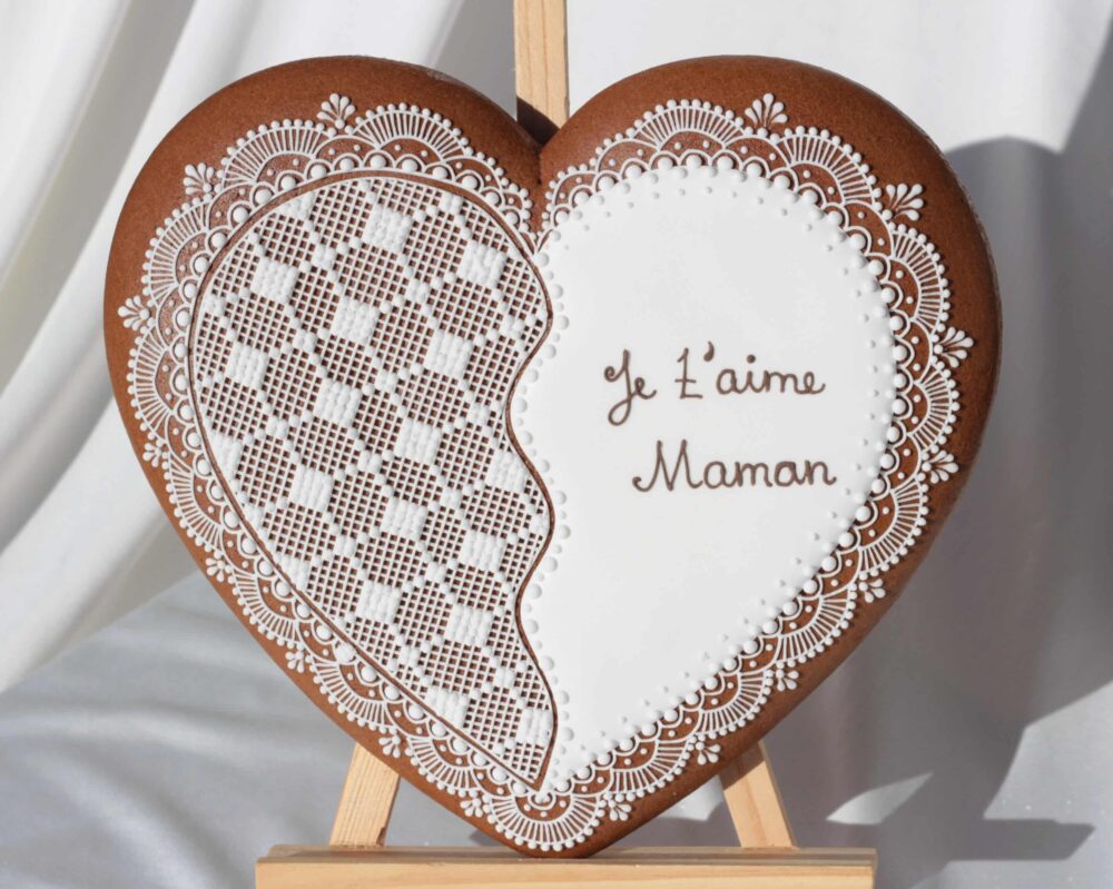 Biscuit en forme de cœur, décoré de motifs en glaçage blanc et portant l'inscription "Je t'aime Maman", posé sur un chevalet.