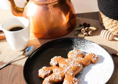 Biscuit décoré en forme de lézard sur une assiette noire, accompagné d'une théière en cuivre, d'une tasse de café et d'épices pour une ambiance conviviale.