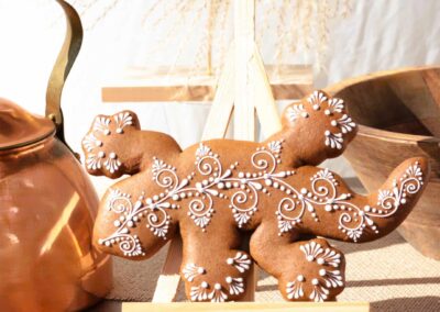 Biscuit décoré en forme de lézard avec des motifs en glaçage blanc, présenté sur un chevalet devant une théière en cuivre.