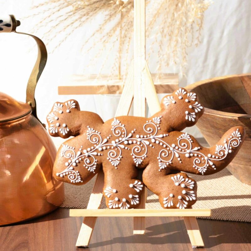 Biscuit décoré en forme de lézard avec des motifs en glaçage blanc, présenté sur un chevalet devant une théière en cuivre.