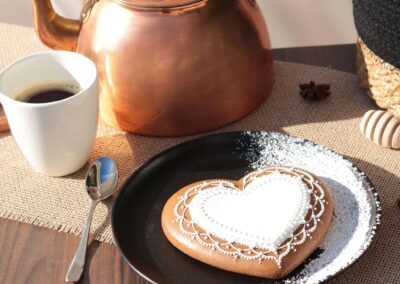 Biscuit en forme de cœur orné de motifs en glaçage blanc, présenté sur une assiette noire avec une théière en cuivre et une tasse de café.