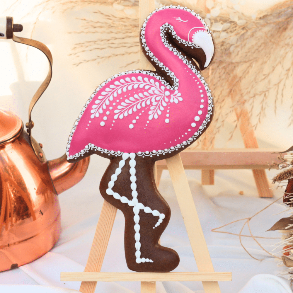 Biscuit en pain d'épices en forme de flamant rose, décoré avec un glaçage rose et blanc aux motifs détaillés, présenté sur un chevalet en bois.